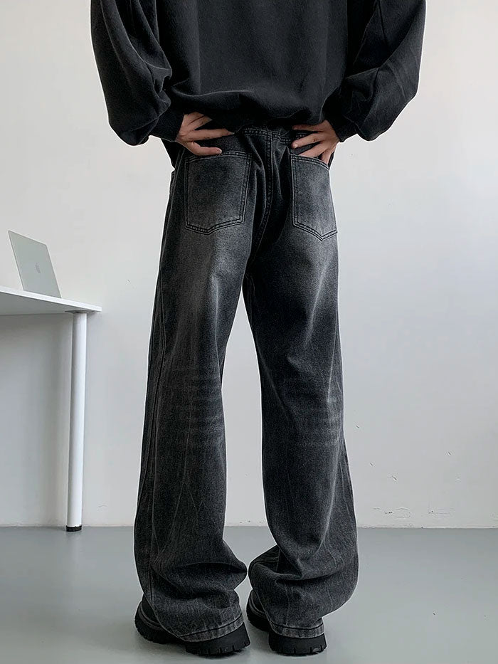 Side view of man in washed black relaxed-fit jeans leaning over white desk in minimalist room.