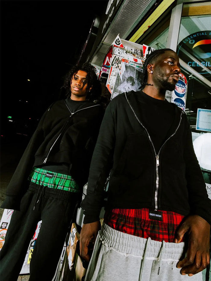 Two models wearing Concrete Mans street-style jogger pants with plaid waistband exposed, night street scene.